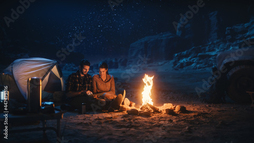 Happy Couple Nature Camping in the Canyon at Night, Use Digital Tablet Computer Sitting by Campfire. Two Traveling people Post Inspirational Traveling Advice and Photos on Internet Social Media.