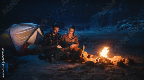 Happy Couple Tent Camping in the Canyon at Night, Use Digital Tablet Computer Sitting by Campfire. Two people Post Inspirational Traveling Advice, Photos on Social Media, Use Internet, do e-Shopping © Gorodenkoff