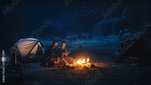 Happy Couple Camping in the Canyon at Night, Use Digital Tablet Computer, Sitting by Campfire. Two people Have Fun, Smile, Post Traveling Photos on Social Media, Watch Funny Videos on Internet