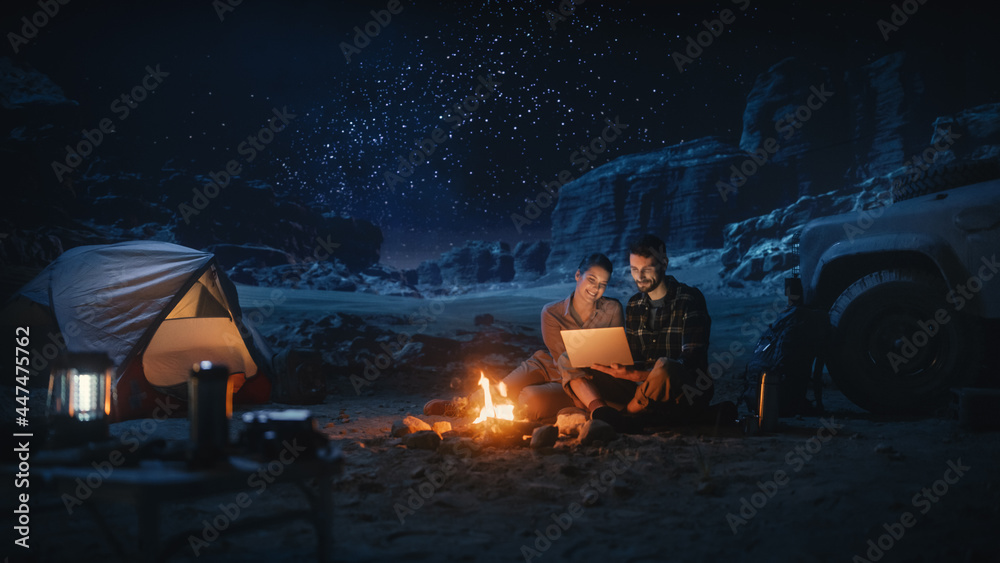 Happy Couple Nature Camping in the Canyon at Night, Use Laptop Computer, Sitting by Campfire. Two Traveling people Have Fun, Smile, Post on Social Media, Watch Funny Video Shows on Streaming Service
