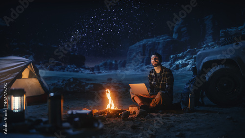 Night Tent Camping in Canyon: Male Traveler Uses Laptop Computer Sitting by Campfire. Man on Digital Remote Work, e-shopping, ecommerce, Using Internet, Social Media Posting on Vacation Trip 