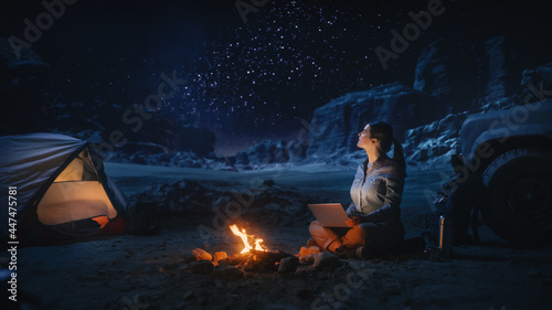 Night Tent Camping in Canyon: Female Traveler Uses Laptop Computer Sitting by Campfire. Woman on Digital Remote Work, e-shopping, ecommerce, Using Internet, Social Media Posting on Vacation Trip 