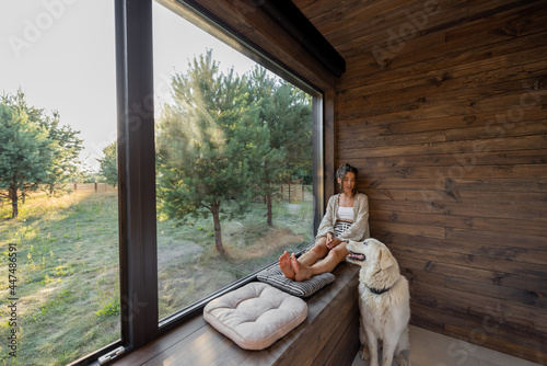 Quadro su tela Young woman resting at beautiful country house or hotel, sitting on the window sill enjoying beautiful view on pine forest with big white dog