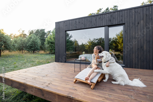 Fototapeta Woman enjoys the nature while sits on sunbed on wooden terrace near the modern house with panoramic windows near pine forest while hugs her pet