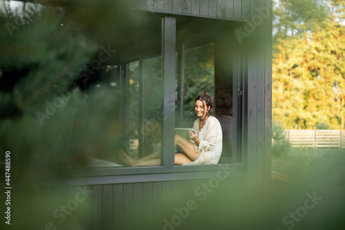 Young woman resting at beautiful country house or hotel, sitting with tablet on the window sill. View from outside. Concept of solitude and recreation on nature. Beautiful destinations for vacation.