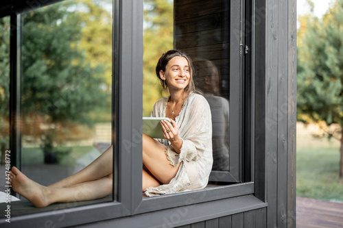 Young cheerful woman enjoy of resting at modern house or hotel, sitting with tablet on the window sill and looking at pine forest. View from outside. Concept of solitude and recreation on nature.