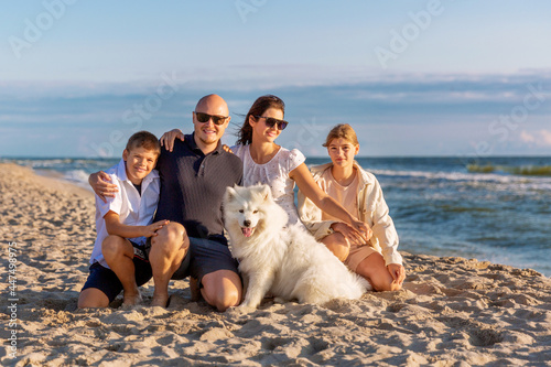 A young happy family with two children and a white husky samoyed dog are sitting hugging on the seashore. Holidays with pets  family relations  parents with children concept
