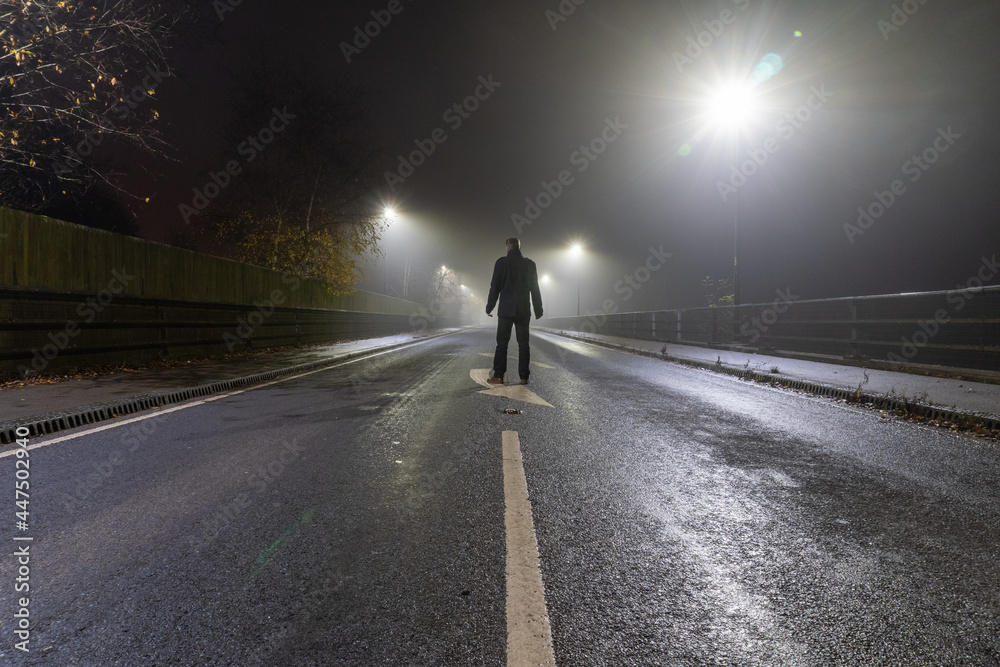A mysterious man standing on a straight road at night on a foggy winters evening. With street lights glowing in the dark.