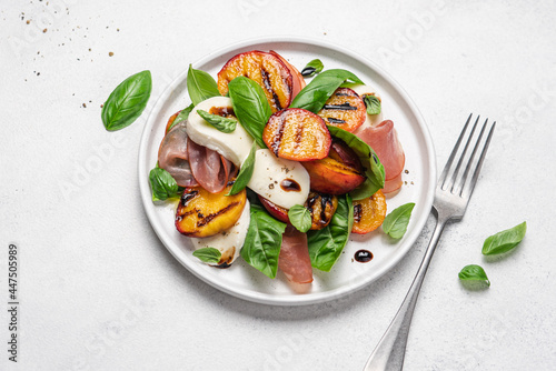 Grilled peach salad with mozzarella, prosciutto ham, basil, olive oil and balsamic. Fresh gourmet salad in plate on white stone background