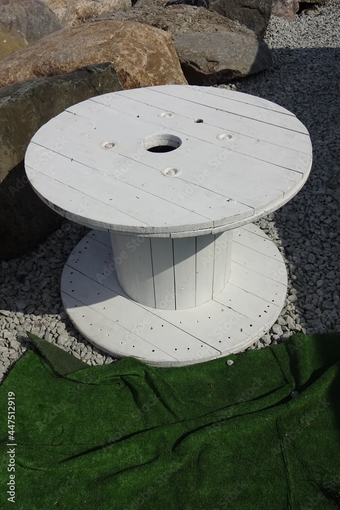 White painted wooden coil for cable or rope. Big stones on the back. Green material in the front. White painted wooden coil for cable or rope. Big stones on the back. Green material in the front.
