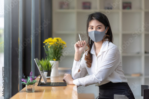 Asian businesswoman sitting at the office holding a stylus with a tablet at the office desk wear a mask to prevent germs. Look at the camera. photo