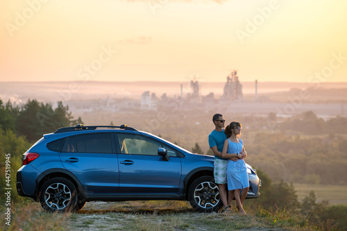 Happy couple standing near their car at sunset. Young man and woman enjoying time together travelling by vehicle. © bilanol