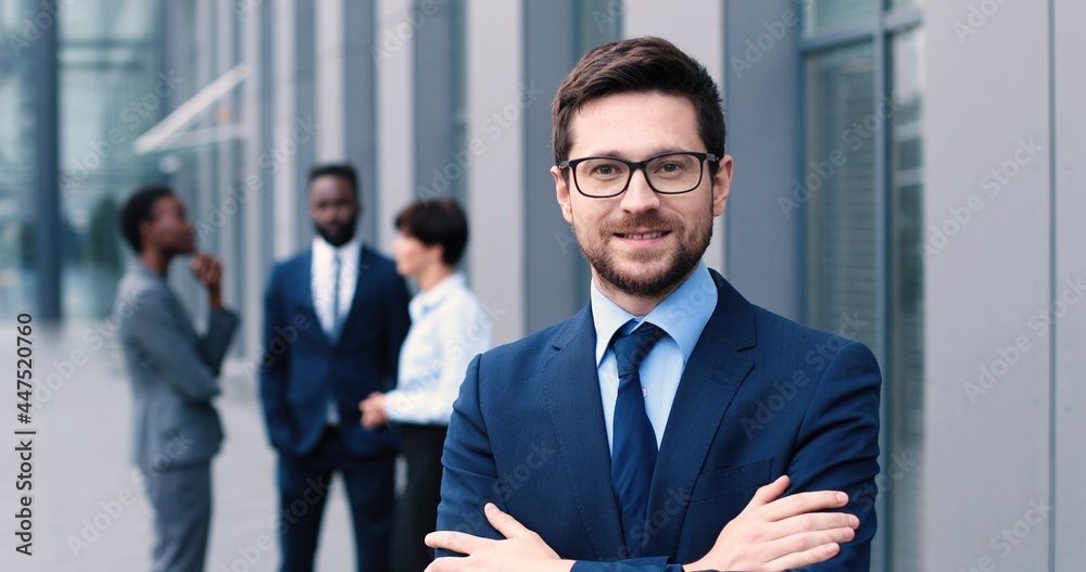 Confident and successful. Waist-up portrait of young elegant caucasian businessman is standing with crossed arms while looking at the camera with slight smile with his colleagues at the background Confident and successful. Waist-up portrait of young elegant caucasian businessman is standing with crossed arms while looking at the camera with slight smile with his colleagues at the background