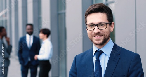 Confident and successful. Waist-up portrait of young elegant caucasian businessman is standing with crossed arms while looking at the camera with slight smile with his colleagues at the background
