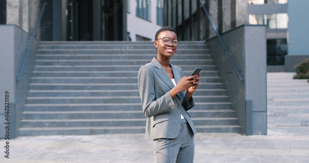 Attractive, adult multiracial woman in chic formal wear standing near the business center. She looking at camera and holding telephone with mail email in hands Attractive, adult multiracial woman in chic formal wear standing near the business center. She looking at camera and holding telephone with mail email in hands