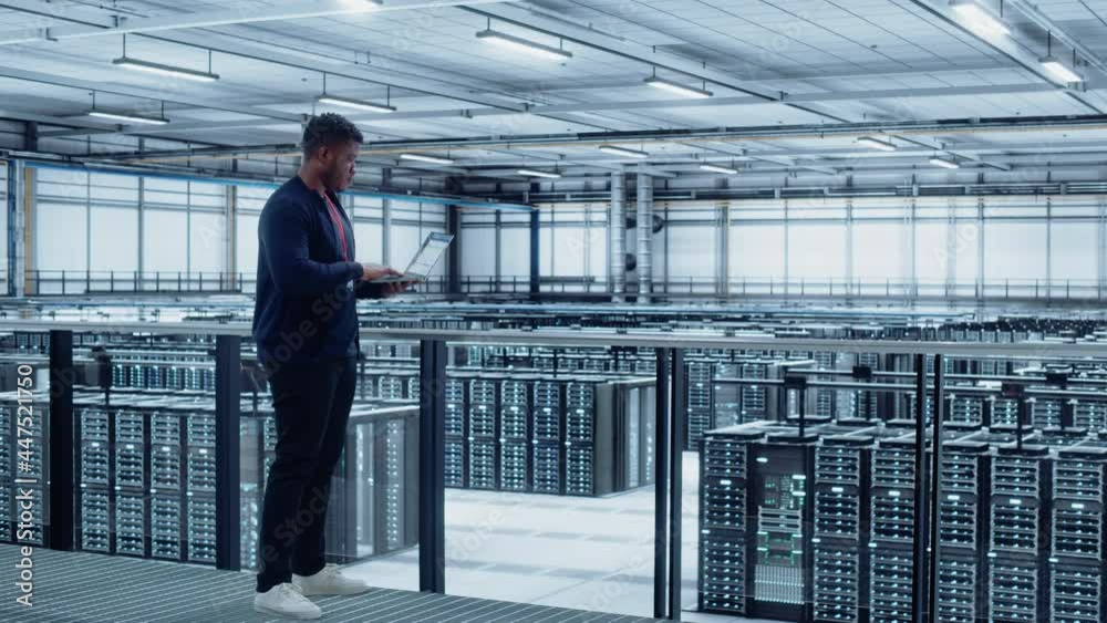 Data Center Engineer Using Laptop Computer. Server Farm Cloud Computing Specialist Facility with African American Male System Administrator Working with Data Protection Network for Cyber Security.
