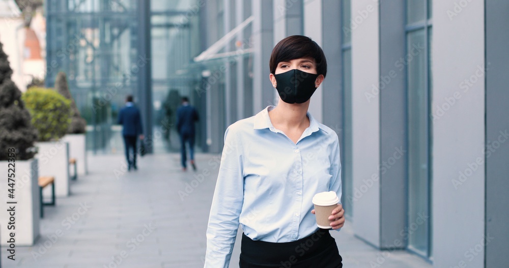 Portrait of beaming young businesswoman wearing protective mask walking through the street and drinking coffee. People and career concept