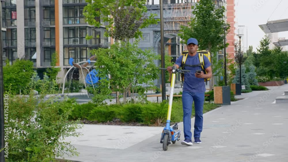 Afro-american man courier food delivery with yellow thermal backpack walks street with electric scooter uses smartphone navigate. Deliveryman worker employee deliver online order client customer Afro-american man courier food delivery with yellow thermal backpack walks street with electric scooter uses smartphone navigate. Deliveryman worker employee deliver online order client customer