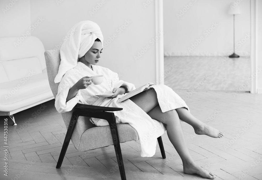 Attractive girl in bathrobe and towel sits on armchair, drinks coffee and reads book. Portrait of beautiful young girl at home in the morning. Black and white - obrazy, fototapety, plakaty 