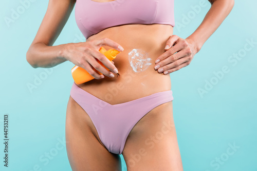 cropped view of woman in swimwear applying sunscreen on body isolated on blue