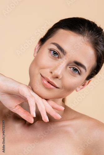 smiling woman with perfect skin touching chin while looking at camera isolated on beige, beauty concept