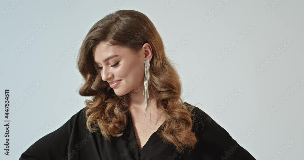 Beautiful young girl with hairstyle and makeup looks into the camera smiling ashamed elegantly dressed beautiful earrings standing on a white background