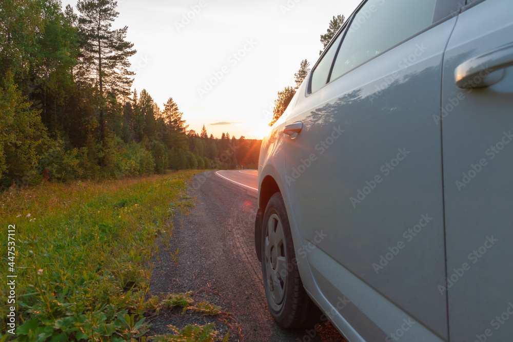 On the side of the road, a part of a white car against the backdrop of a sunset in the forest. Green trees. Selective focus. Close-up