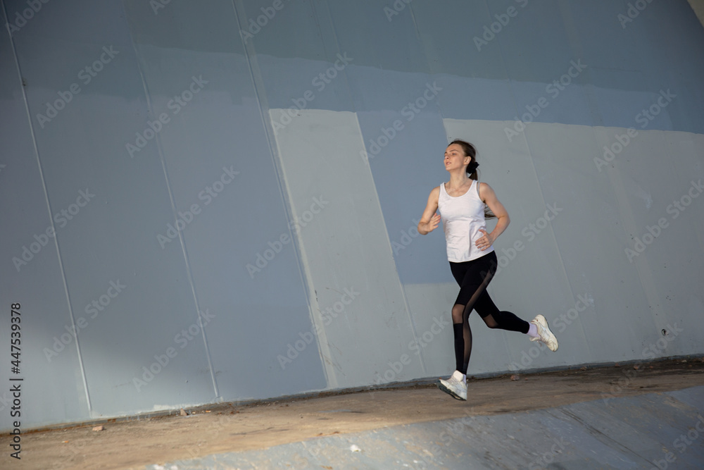 A young woman in a white T-shirt and black pants runs under the bridge in the evening. Running trainings in the city. Preparing for the competition. A young woman in a white T-shirt and black pants runs under the bridge in the evening. Running trainings in the city. Preparing for the competition.