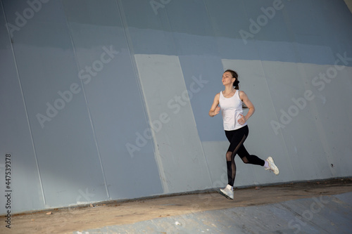 A young woman in a white T-shirt and black pants runs under the bridge in the evening. Running trainings in the city. Preparing for the competition. © Алексей Васильев