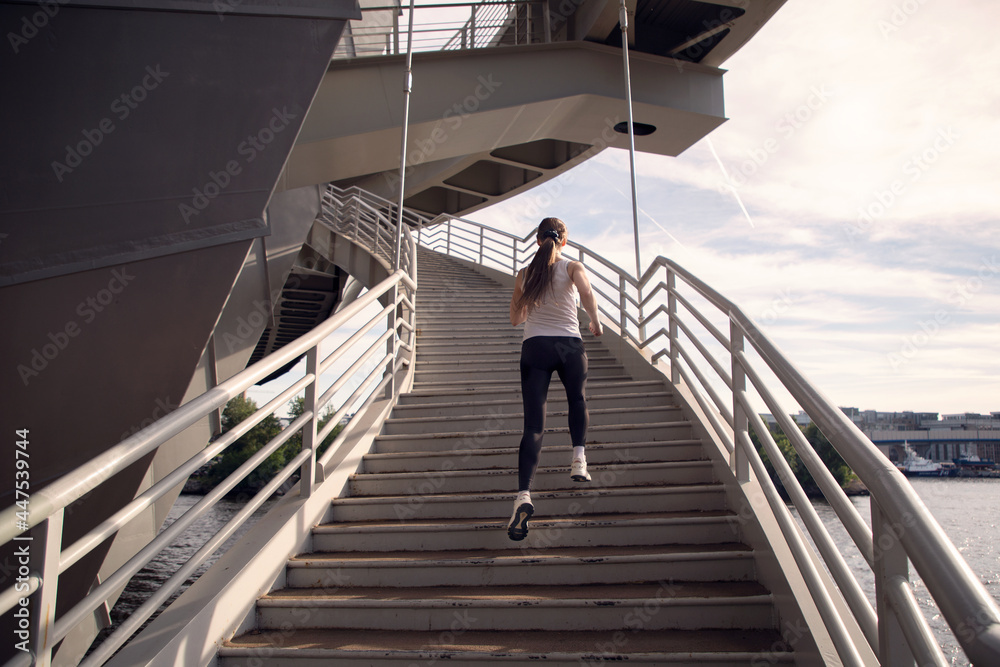 A young woman runs up the stairs of the bridge. Power training. Running in the evening in the city