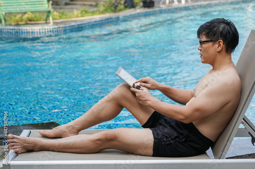 Asian man in swimwear sitting by swimming pool working on digital tablet. work remotely in summer season.