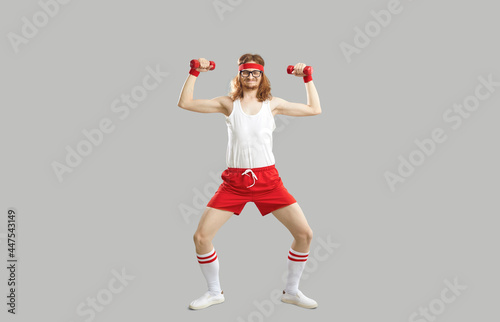 Full body skinny nerd with funny face doing exercise with dumbbells isolated on gray background. Full length hilarious smiling man in sweatband, white tank top and red shorts enjoying sports workout photo