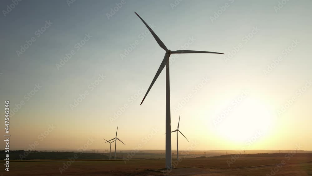 Large wind turbines with blades in field aerial view bright orange sunset blue sky wind park slow motion drone turn. Silhouettes windmills, large orange sun disc summer lens flare. Alternative energy Large wind turbines with blades in field aerial view bright orange sunset blue sky wind park slow motion drone turn. Silhouettes windmills, large orange sun disc summer lens flare. Alternative energy