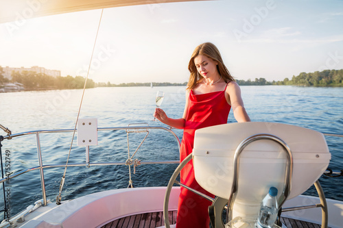 The elegant attractive woman in a long red dress holding a glass of champagne standing on a yacht and looking for river sunset view