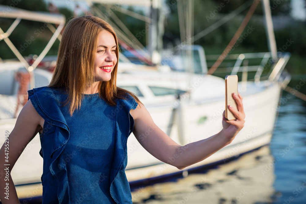 Attractive woman taking selfie photo on smartphone for her blog while standing on the river marina. Tourism and travel concept.