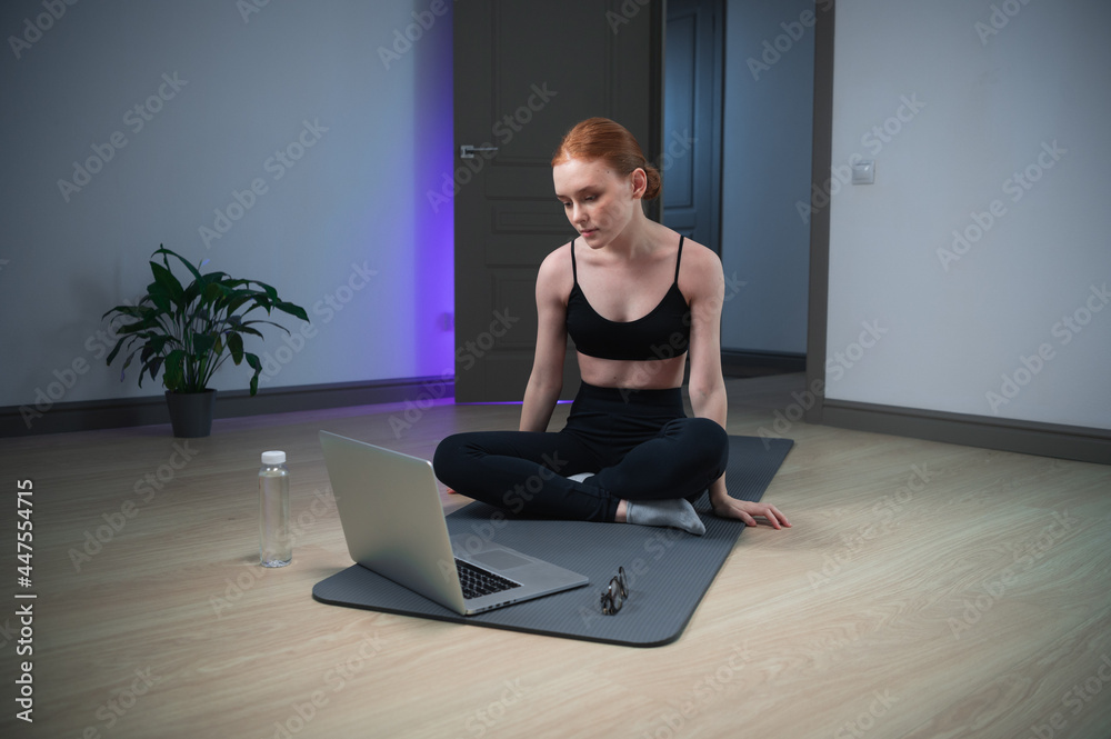 A young mother wants to tone up her body during maternity leave, she sits on the floor of the house on a gym mat and prepares for exercises in the app on her laptop A young mother wants to tone up her body during maternity leave, she sits on the floor of the house on a gym mat and prepares for exercises in the app on her laptop