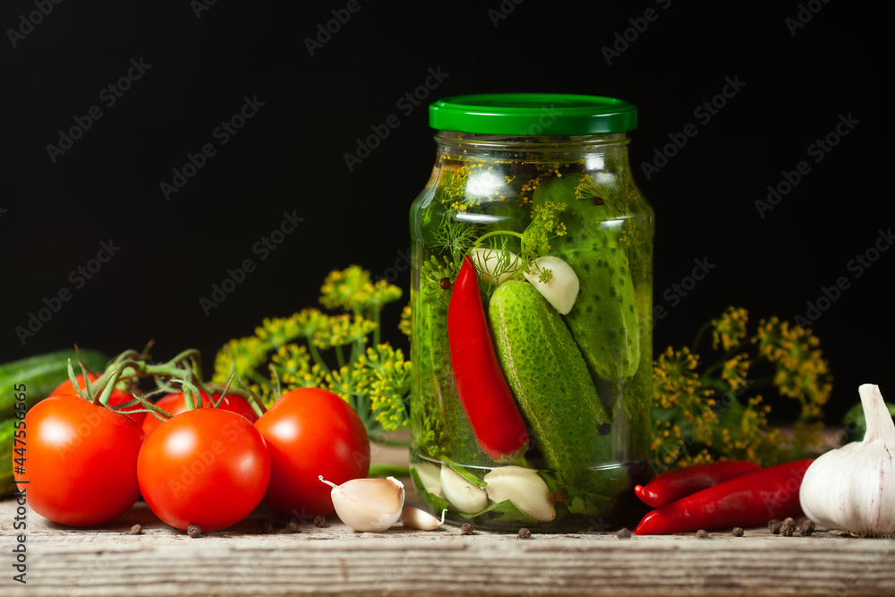 Pickled cucumbers in glass jars and spices and vegetables for preparation of pickles on black background Pickled cucumbers in glass jars and spices and vegetables for preparation of pickles on black background