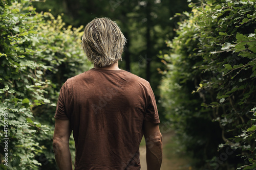 Blonde man in a brown t-shirt on a footpath with a hedge on eith