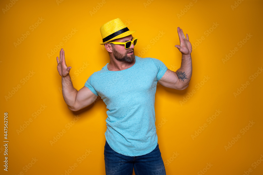 Happy male in blue shirt, sunglasses and yellow hat celebrating success standing on yellow background. 