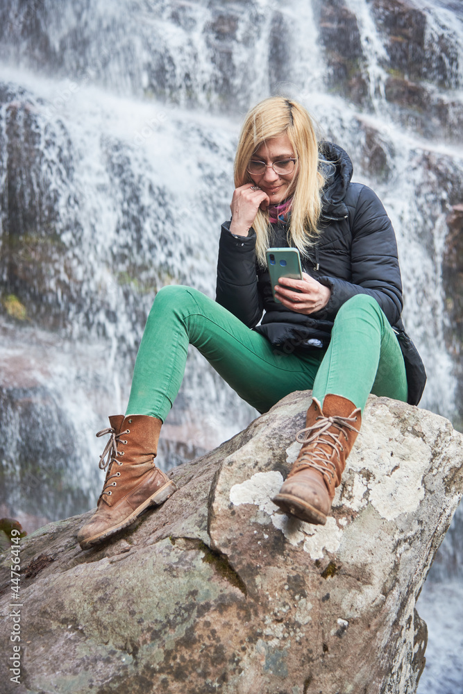 Young woman sitting on a rock in front of a big waterfall, chatting on her phone to her friend, reporting news of her adventure Young woman sitting on a rock in front of a big waterfall, chatting on her phone to her friend, reporting news of her adventure