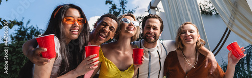 Positive interracial friends with plastic cups hugging outdoors, banner