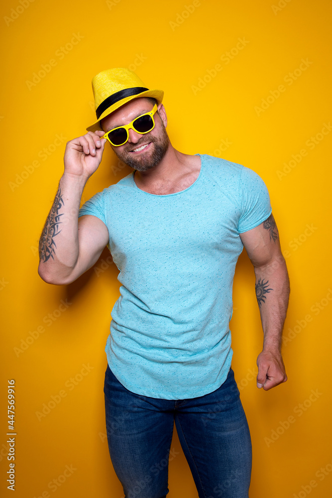 Lively young bearded male in bright blue shirt, jeans and yellow stylish hat standing in studio touching yellow sunglasses with hand and looking at camera 