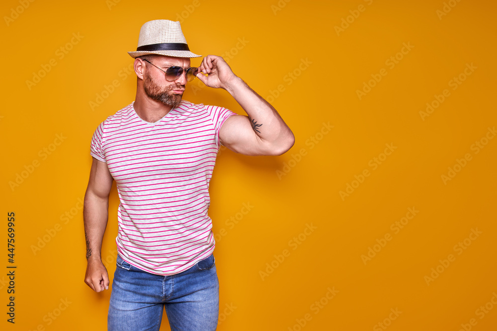Muscular bearded male in shirt, jeans and stylish hat, sunglasses standing in studio over yellow background