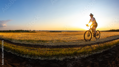 The cyclist rides a bike on the road near the field against the backdrop of the setting sun. Outdoor sports. Healthy lifestyle.