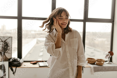 Pretty brunette woman in white blouse smiles sincerely with closed eyes. Attractive girl poses near window at home.