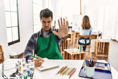Young artist man at art studio doing stop gesture with hands palms, angry and frustration expression