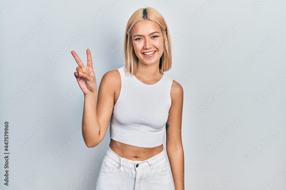 Beautiful blonde woman wearing casual style with sleeveless shirt showing and pointing up with fingers number two while smiling confident and happy.