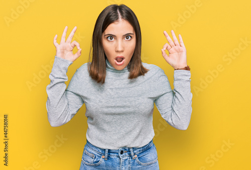 Young beautiful woman wearing casual turtleneck sweater looking surprised and shocked doing ok approval symbol with fingers. crazy expression