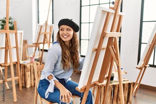 Young hispanic artist woman painting on canvas at art studio looking away to side with smile on face, natural expression. laughing confident.