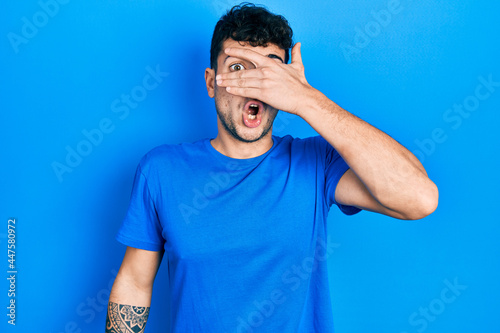 Young hispanic man wearing casual blue t shirt peeking in shock covering face and eyes with hand, looking through fingers with embarrassed expression.
